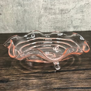 May include: A pink glass footed bowl with a scalloped edge. The bowl has a clear glass base and three feet.