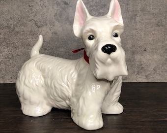 Peut inclure: Figurines en céramique blanche d'un chien terrier écossais. Le chien est assis, la tête légèrement inclinée sur le côté. Il porte un ruban rouge autour du cou et un charme argenté suspendu à son collier.