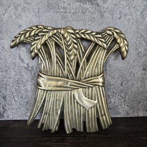 Pode incluir: Uma decoração de parede de metal dourado em forma de espiga de trigo com uma fita amarrada ao redor do centro. A espiga é feita de metal texturizado e detalhado.