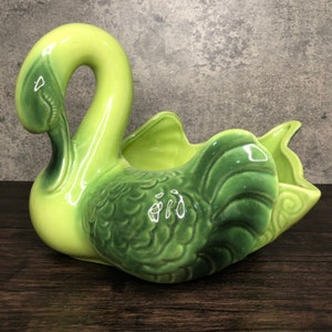 May include: A green ceramic swan planter with a textured design. The swan's head is turned to the side and its wings are spread out. The planter has a large opening in the centre for planting.