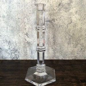 May include: A clear glass candle holder with a hexagonal base and a tall, cylindrical body. The holder has a decorative, geometric design.