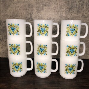 May include: Set of six white ceramic mugs with a blue and green floral pattern. The mugs are stacked in three pairs.
