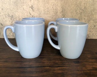 May include: Four light blue ceramic mugs with rounded handles. The mugs are arranged in a row on a dark brown wooden surface.