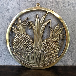 May include: A brass wall hanging featuring two pineapples with detailed carvings. The pineapples are surrounded by leaves and have a textured surface.