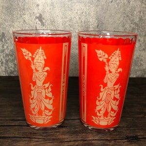 May include: Two clear drinking glasses with a vibrant orange design. Each glass features a cream-colored illustration of a figure in traditional garb. The glasses are ideal for serving beverages and add a touch of vintage charm to any table setting.