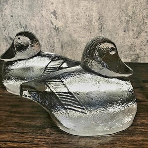 Puede incluir: Dos figuras de pato de cerámica con acabado plateado metálico. Los patos tienen picos y ojos negros, y las alas están detalladas con líneas grabadas. Las figuras se exhiben sobre una superficie de madera contra un fondo gris texturizado.