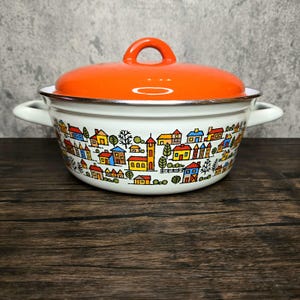 Könnte beinhalten: Ein weißer Emailletopf mit einem orangefarbenen Deckel und einem bunten Stadtmotiv. Der Topf hat zwei Griffe und einen kleinen Knopf auf dem Deckel.