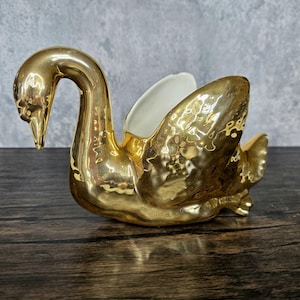 May include: A gold swan-shaped ceramic planter with a white interior. The planter has a textured, hammered finish.