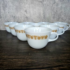 May include: Set of ten white ceramic coffee mugs with a gold floral pattern around the rim.
