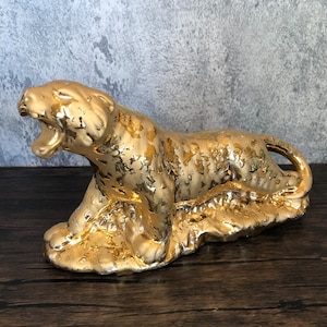 Könnte beinhalten: Eine goldene Keramikfigur eines brüllenden Leoparden, der auf dem Bauch liegt. Die Figur ist mit einem strukturierten, gefleckten Muster verziert.