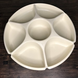 Op de afbeelding: Crèmekleurige, verdeelde serveerschaal met een centrale dipschaal en zes omliggende compartimenten. De ronde schaal is gemaakt van een glad, glanzend materiaal, perfect voor het serveren van snacks of hapjes. Het ontwerp is eenvoudig en functioneel.