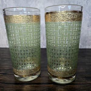 May include: Two tall, clear glass tumblers with a green and gold geometric pattern. The glasses have a gold band around the top and bottom.