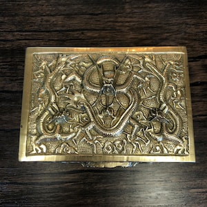 Puede incluir: Una caja de latón con un diseño intrincado de dragones y nubes. La caja es rectangular y tiene una tapa con bisagras.