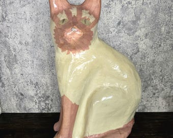 May include: A ceramic cat figurine with pink and white paint. The cat is sitting with its head tilted slightly to the side.