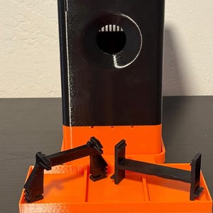 May include: A 3D-printed black and orange object with a rectangular base and a tall, rectangular tower. The tower has a circular opening with vertical lines inside. Two black, angular pieces sit on an orange tray.