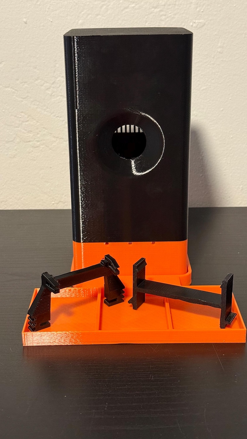 May include: A 3D-printed black and orange object with a rectangular base and a tall, rectangular tower. The tower has a circular opening with vertical lines inside. Two black, angular pieces sit on an orange tray.