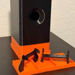 May include: A black and orange 3D-printed object with a rectangular base and a cylindrical hole. The object has a black upper section and an orange lower section. Several black, geometric components are placed on an orange tray.