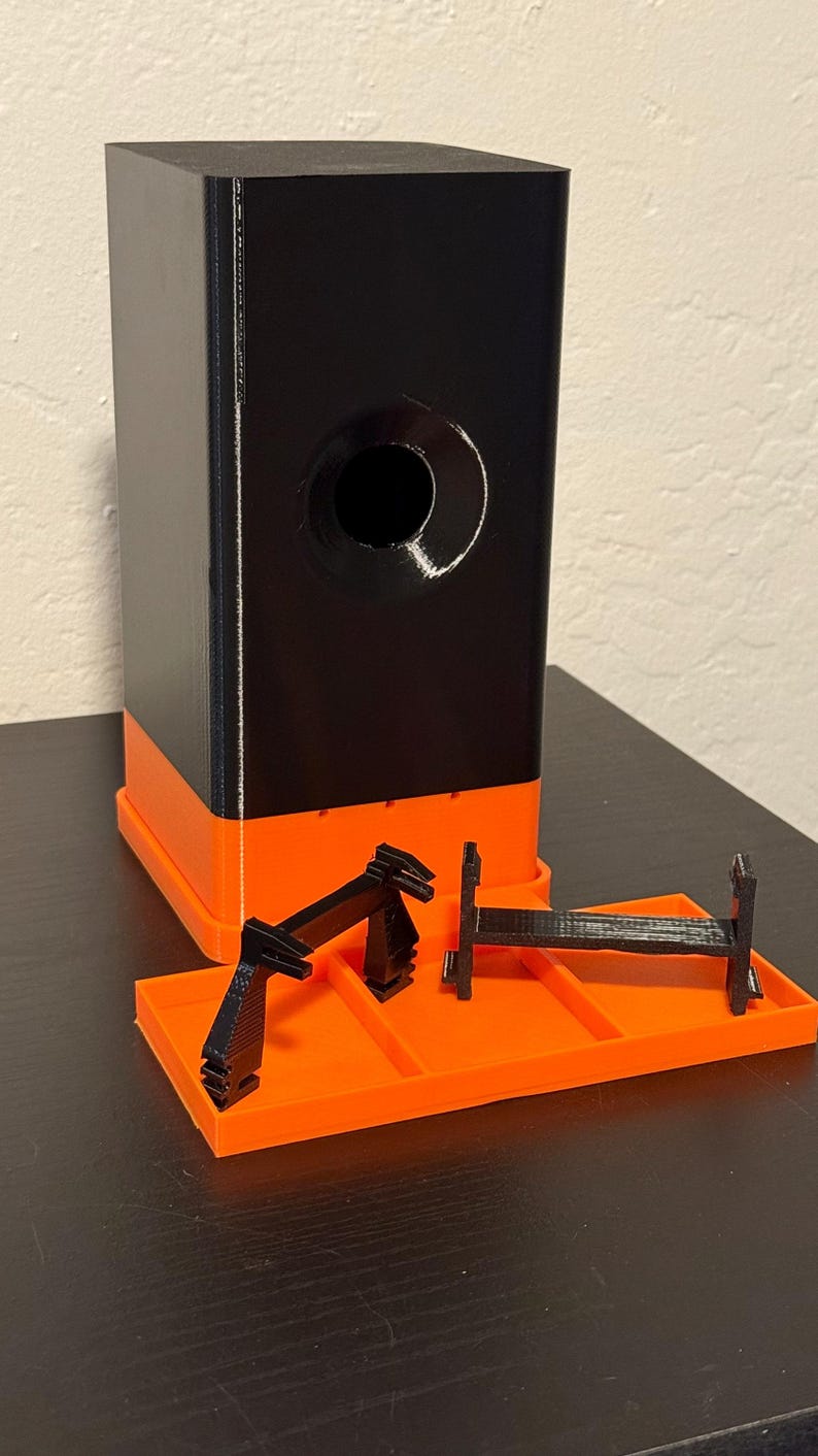 May include: A black and orange 3D-printed object with a rectangular base and a cylindrical hole. The object has a black upper section and an orange lower section. Several black, geometric components are placed on an orange tray.