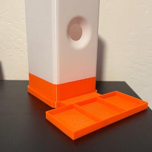 May include: A white and orange 3D-printed dispenser with a rectangular shape and a circular opening. The dispenser has an orange base and a matching tray. The tray is divided into three sections.