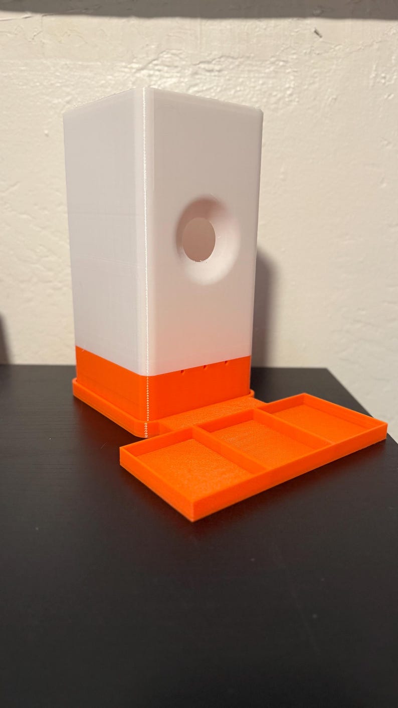 May include: A white and orange 3D-printed dispenser with a rectangular shape and a circular opening. The dispenser has an orange base and a matching tray. The tray is divided into three sections.