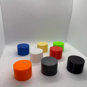 May include: A collection of eight cylindrical objects in various colors: orange, blue, yellow, white, red, green, gray, and black. Each cylinder has a flat top and bottom, and a smooth, matte finish. The objects are arranged on a white surface.