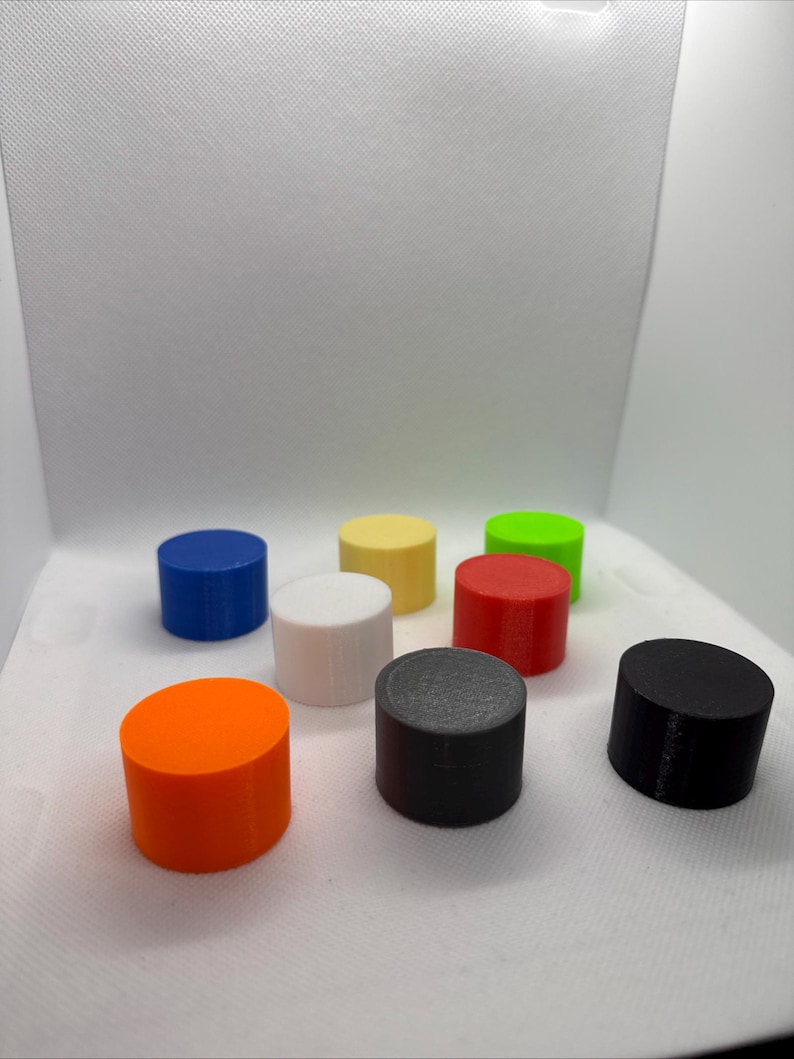 May include: A collection of eight cylindrical objects in various colors: orange, blue, yellow, white, red, green, gray, and black. Each cylinder has a flat top and bottom, and a smooth, matte finish. The objects are arranged on a white surface.