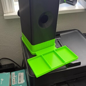 May include: A 3D-printed black and neon green device with a rectangular base and a tray. The device has a cylindrical opening and a tray with three compartments. It is sitting on top of a black printer.