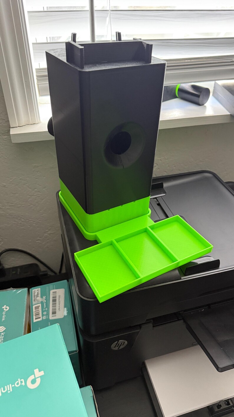 May include: A 3D-printed black and neon green device with a rectangular base and a tray. The device has a cylindrical opening and a tray with three compartments. It is sitting on top of a black printer.