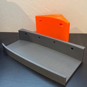Puede incluir: Una estantería gris, impresa en 3D, con un borde frontal curvo y agujeros de montaje. Detrás hay una pieza triangular naranja con dos agujeros. Ambos artículos están sobre una superficie oscura.