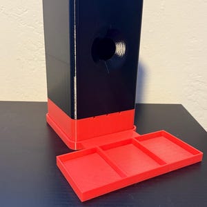 May include: A black and red 3D-printed dispenser with a rectangular base and a tray with three compartments. The dispenser has a large circular opening and a rectangular top.