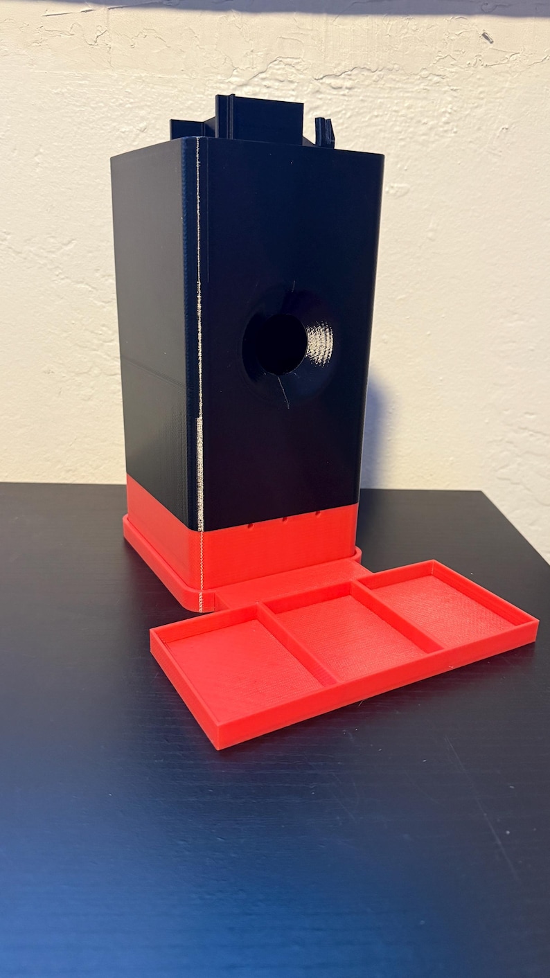 May include: A black and red 3D-printed dispenser with a rectangular base and a tray with three compartments. The dispenser has a large circular opening and a rectangular top.