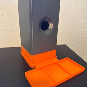 May include: A 3D-printed, two-tone storage container. The main body is gray with an orange base and a circular opening. An attached orange tray with three compartments extends from the base.