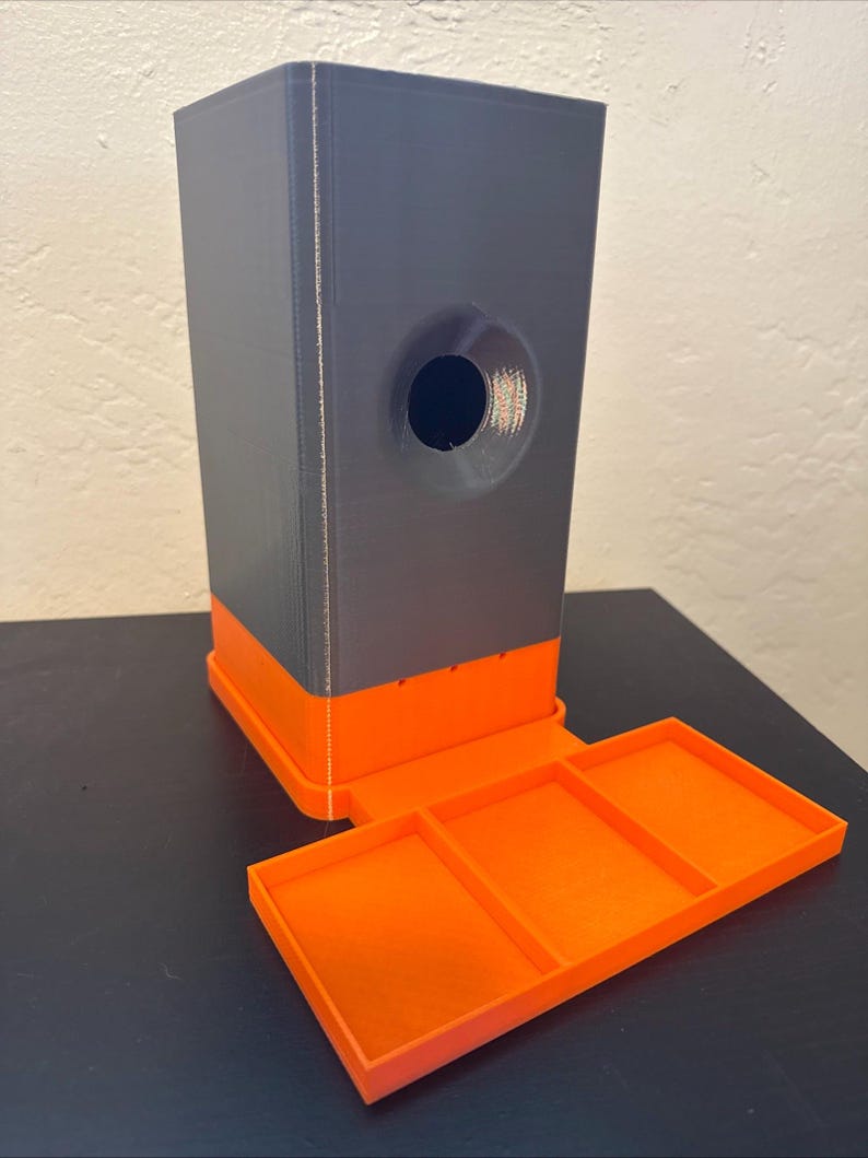 May include: A 3D-printed, two-tone storage container. The main body is gray with an orange base and a circular opening. An attached orange tray with three compartments extends from the base.