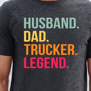 May include: Dark gray t-shirt with the words "HUSBAND. DAD. TRUCKER. LEGEND." in teal, yellow, orange, and red block letters. The shirt is short-sleeved and has a crew neck.