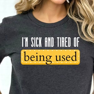 May include: Dark gray t-shirt with the text "I'M SICK AND TIRED OF being used" in white and black. The text "being used" is in a yellow rectangle with a stitched border. The shirt is short-sleeved.