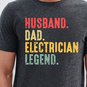 May include: Dark gray t-shirt with the words "HUSBAND. DAD. ELECTRICIAN. LEGEND." in red, yellow, and green block letters. The shirt is a crew neck and has short sleeves.