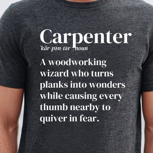 Puede incluir: Camiseta gris oscuro con la palabra "Carpenter" en blanco, junto con una definición. La definición describe a un mago de la carpintería. La camiseta es de cuello redondo y tiene mangas cortas.