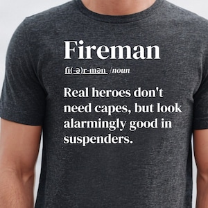 May include: Dark gray t-shirt with the word "Fireman" and its pronunciation in white text. The shirt also features the quote: "Real heroes don't need capes, but look alarmingly good in suspenders."