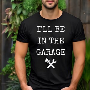 以下が含まれることがあります： 白い文字で「I'LL BE IN THE GARAGE」とレンチとハンマーのグラフィックがプリントされた黒いクルーネックTシャツ。柔らかい素材で作られています。