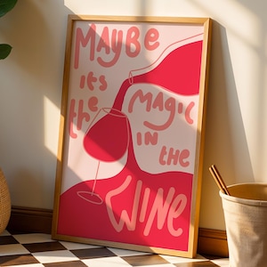 May include: A framed art print with the text "Maybe it's the magic in the wine" in pink and red. The artwork depicts a wine bottle pouring into a wine glass. The frame is a light wood color.