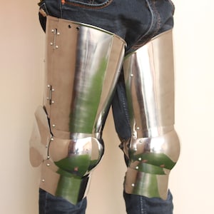 Puede incluir: Armadura de piernas de acero pulido, con placas articuladas para rodillas y muslos. La armadura está remachada y refleja el entorno. El diseño recuerda a la armadura de caballero medieval.