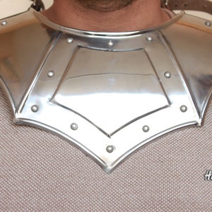 Könnte beinhalten: Ein silberner Nackenschutz aus Metall mit einem spitzen, gewellten Rand und einem zentralen, rautenförmigen Feld. Der Schutz ist mit Nieten befestigt.
