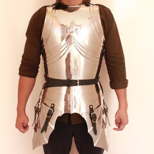 May include: A silver-coloured metal breastplate and tassets with a decorative fleur-de-lis design. The armour is worn over a brown shirt and a black belt.
