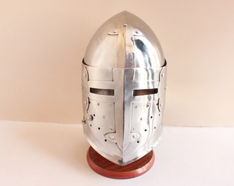 May include: A silver helmet with a cross design on the front and a visor. The helmet is on a wooden stand.