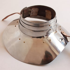 Könnte beinhalten: Ein silberner Nackenschutz aus Metall mit einem Lederriemen und Messingnieten. Der Schutz ist wie ein breiter Kragen geformt und hat eine abgerundete Kante.