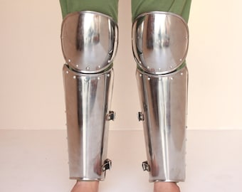 May include: A pair of silver knee-high leg armor with buckles. The armor is made of metal and has a shiny finish.