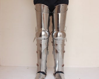 May include: A pair of silver-colored metal leg armor with knee pads and straps. The armor is worn over black pants.