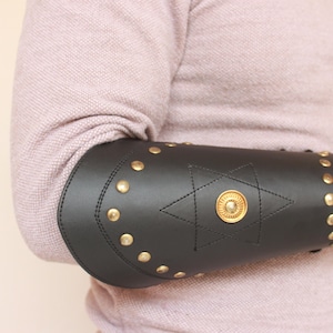 以下が含まれることがあります： 金色のスタッズと星形のデザインが施された黒い革製の腕カバー。腕カバーの中央の星には、金色の円形のアクセントが施されています。