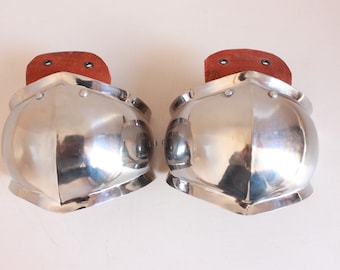 May include: A pair of silver metal elbow pads with brown leather straps. The pads are rounded and have a slightly pointed shape at the top.