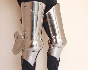 Puede incluir: Un par de armaduras de rodilla plateadas con correas de cuero. La armadura está hecha de placas de metal y tiene una rodillera redondeada. La armadura se lleva sobre pantalones negros.